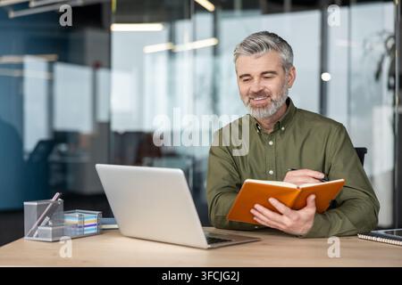 Selbstbewusster Senior-Manager arbeitet in einem modernen Büro, lächelt, während er seinen Laptop beobachtet und wichtige Notizen in einem offenen Planer schreibt, konzentriert auf Strategie und Planung für den Geschäftserfolg Stockfoto