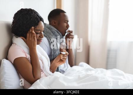 Krankes schwarzes Paar in Schals gehüllt, das zusammen im Bett im Schlafzimmer sitzt und Fieber und Niesnasen misst. Junge afrikanische Familie von zwei wurde krank, bleiben Stockfoto