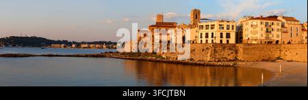 Panoramische Ansicht der Altstadt von Antibes, Frankreich, zur Goldenen Stunde mit mittelalterlichen Türmen, Steinmauern und Häusern am Wasser, die über einer ruhigen Bucht und sandigem Ufer leuchten Stockfoto