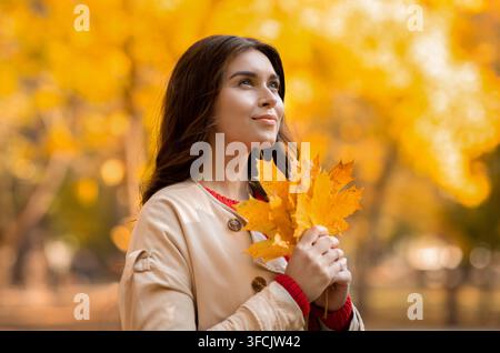 Verträumte junge Frau im stilvollen Herbstmantel mit gelben Blättern Im Park Stockfoto