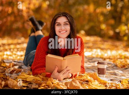 Lächelnde junge Frau mit Buch und Kaffee zum liegen gehen Auf Decke zwischen herbstlichen Blättern und warmen Herbst genießen Tag Stockfoto