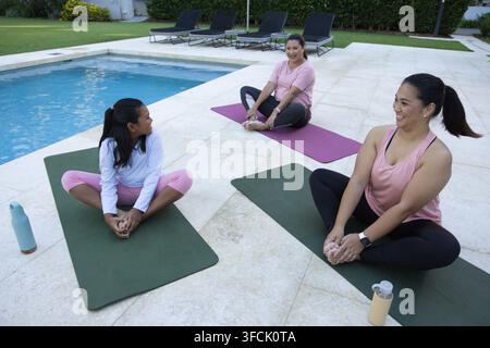 Verschiedene weibliche Freunde, einschließlich Senioren und Jugend, sitzen auf einem gefliesten Pooldeck auf Yogamatten und lächeln Stockfoto