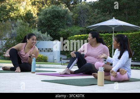 Verschiedene weibliche Familie sitzt, plaudert in Sportkleidung auf Yogamatten am Pooldeck mit Wasserflaschen Stockfoto