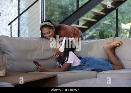 Verschiedene Mutter und Kind, die sich auf ein beiges Sofa lehnen, das Kind hält ein Smartphone und trägt Kopfhörer Stockfoto