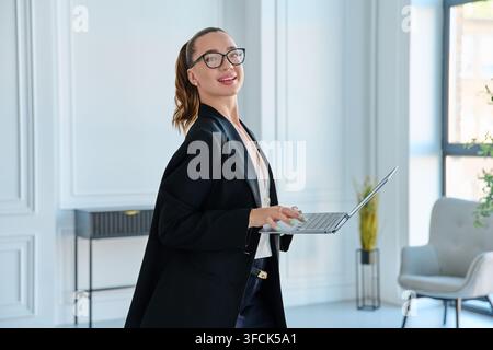 Lächelnde Geschäftsfrau mit Brille, die einen Laptop hält, im modernen Büro stehend Stockfoto