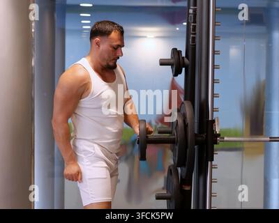 Ein muskulöser, bärtiger Mann in vollständig weißer Sportkleidung (geripptes Tanktop und Shorts) wird in einer Dreiviertel-Rückansicht aufgenommen. Er lädt aktiv eine große, b Stockfoto
