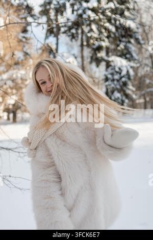 Glückliche blonde Frau mit langen Haaren, die sich in einem verschneiten Wald dreht, verspielter Winter-Lebensstil und warmes Mode-Porträt im Freien. Vertikates Format. Stockfoto