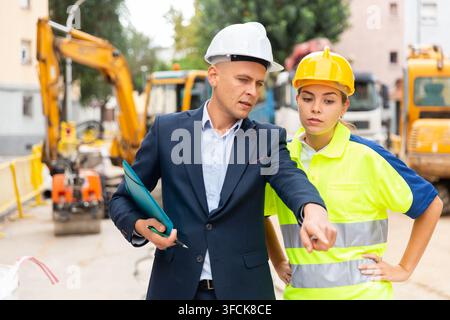 Vorarbeiter in Anzug und weißem Schutzhelm plant die Arbeit mit einer weiblichen Angestellten in Uniform, während sie auf der Baustelle stehen. Stockfoto