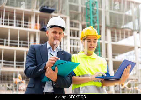 Ein Ingenieur, der mit einer jungen Arbeiterin auf einer Baustelle steht, überwacht den Bauplan auf einem Laptop Stockfoto