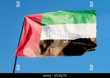 Flagge der Vereinigten Arabischen Emirate. Fahne weht im Wind vor einem klaren blauen Himmel, flattert an einer Fahnenstange im Freien. Heller Sonnentag, Stofftextur Stockfoto