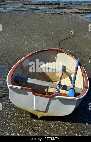 Ein kleines, verwittertes Fiberglas- oder Plastikdingi liegt verlassen auf einer sandigen Gezeitenfläche bei Ebbe, eine ruhige Küstenszene. Stockfoto
