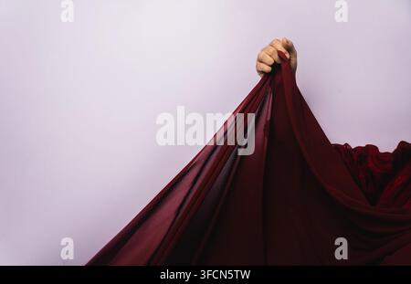 Hand, die grauen Stoff aus Chiffon in Maroon gegen grauen Hintergrund hält und hebt Stockfoto