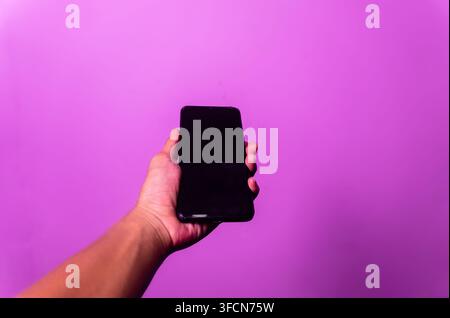 Hand, die ein schwarzes Smartphone mit leerem Bildschirm vor lila Hintergrund hält Stockfoto
