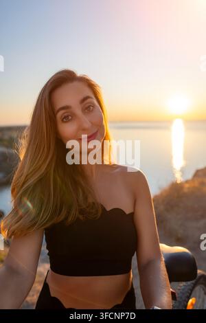 Frau Sonnenuntergang Ozean Fröhliche junge Frau mit langen braunen Haaren genießt die goldene Stunde auf einer malerischen felsigen Klippe während eines schönen Sommerabends Stockfoto