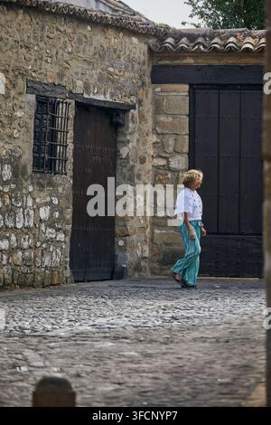 Frau geht an verwitterten Steinbauten mit vergitterten Fenstern, dunklen Holztüren und Ziegeldächern in der Altstadt von Baeza, Spanien, vorbei. Stockfoto
