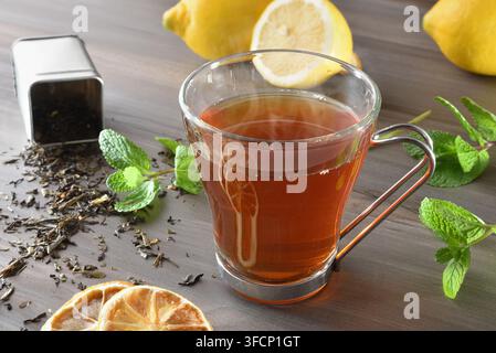 Schwarze Teeinfusion in einer Glas Tasse und ein Behälter voller getrockneter Teeblätter auf einem Holztisch mit Minze und Zitrone. Erhöhte Ansicht. Stockfoto