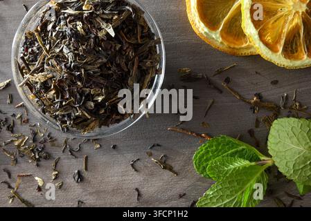 Gläschene Schale gefüllt mit getrockneten Teeblättern auf einem dunklen Holztisch mit dehydrierten Zitronenscheiben und frischen Minzblättern. Top-Down-Ansicht. Stockfoto
