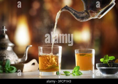Details des Einschenkens von marokkanischem Tee aus einer Metallteekanne in ein Glas auf einem Holztisch mit einer Schale getrockneten Blatttees, verziert mit Minzblättern in einem Teeraum. Stockfoto
