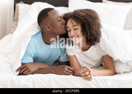 Glücklich junge gut aussehende afroamerikanische Mann küssen Frau auf der Stirn, liegen auf dem Bett Stockfoto