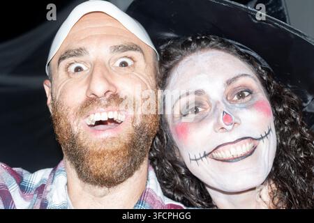 Nahaufnahme-Porträt von zwei Freunden, die eine weiße Maske und eine vom Schädel inspirierte Gesichtsbemalung tragen, während sie zusammen bei einer Halloween-Feier posieren. Stockfoto