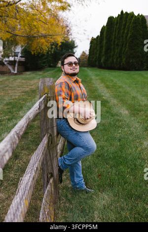 Junger Mann in kariertem Hemd hält einen Cowboyhut an einem Zaun Stockfoto