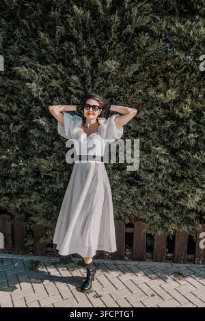 Frau in weißem Kleid im Garten, posiert elegant vor einer grünen Hecke, während sie eine stylische Sonnenbrille und einen schwarzen Hut trägt Stockfoto
