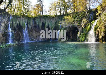Ruhiger türkisfarbener See, der von mehreren kleinen Wasserfällen gespeist wird, die über eine moosbedeckte Kalksteinklippe stürzen, umgeben von üppigem grünen Wald. Stockfoto