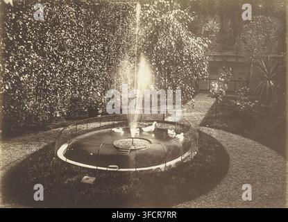 Teich mit Brunnen und Enten in einem Garten, Fotografie, Fotograf: zugeschrieben Richard Tepe, Niederlande, ca. 1900 - ca. 1930, Zugeschrieben, Papier, Salzpapierdruck, Albumen-Druck, Salzdruck auf Fotopapier, Höhe 165 mm x Breite 225 mm., Ein kreisrunder Steinbrunnen steht im mittleren Vordergrund mit einem vertikalen Wasserstrahl, der aus seiner Mitte aufsteigt, und einem umrandenden ringförmigen Metallnetz; mehrere weiße Enten schwimmen auf der Wasseroberfläche; ein geschwungener Rasenrand umgibt den Brunnen und ein Kiesweg verläuft um ihn herum, vorbei an Topfpflanzen und kleinen Sträuchern; dichte Laubhedges und hohe Bäume füllen Stockfoto