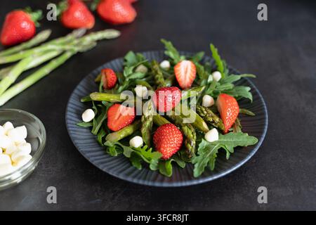 Gesunder Frühlingssalat mit Spargel, Erdbeeren, Rucola und Mozzarellaperlen, serviert in einer dunklen Schale Stockfoto