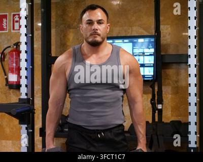 Vorderansicht-Porträt eines selbstbewussten sportlichen Mannes, der aufrecht in einem Hantelbankbereich im Fitnessstudio steht und Kurzhanteln an seinen Seiten hält. Verschwitzte Haut und ein Stockfoto