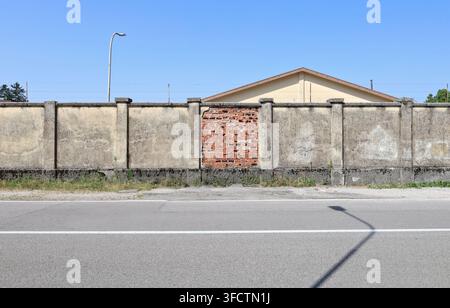 Grunge-Grenzmauer mit der mittleren abgeblätterten Partie, bei der Ziegel freigelegt sind. Stacheldraht oben. Dach, Himmel und Straßenlaterne dahinter, Straße davor. Stockfoto