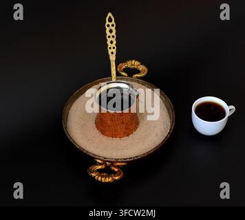 Kupfertürke mit heißem Getränk in der Schale mit Sand und Tasse schwarzem Kaffee auf schwarzem Hintergrund. Oberansicht, Flat Lay. Stockfoto