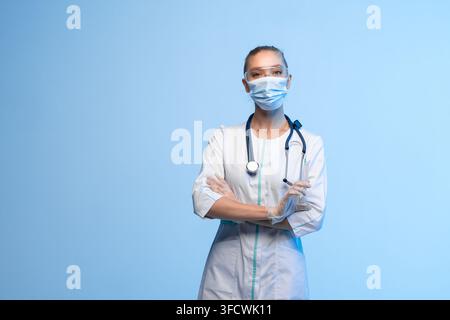 Medizinischer Mitarbeiter in persönlicher Schutzausrüstung, schaut in die Kamera und hält eine Spritze vor blauem Hintergrund Stockfoto