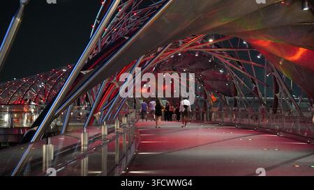 Menschen gehen über die beleuchtete Helixbrücke bei Nacht.Fußgänger überqueren eine moderne Fußgängerbrücke mit einer markanten spiralförmigen Stahlkonstruktion, beleuchtet Stockfoto