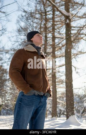 Junger, gutaussehender Mann in brauner Jacke und schwarzem Hut, der unter schneebedeckten Bäumen im hellen Wintersonnenlicht nach oben schaut. Outdoor-Lebensstil und saisonales Wohlbefinden Stockfoto