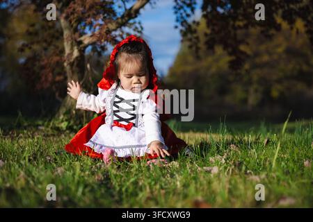 Kleinkind in rotem Kapuzenkostüm, das auf dem Gras für Halloween sitzt Stockfoto