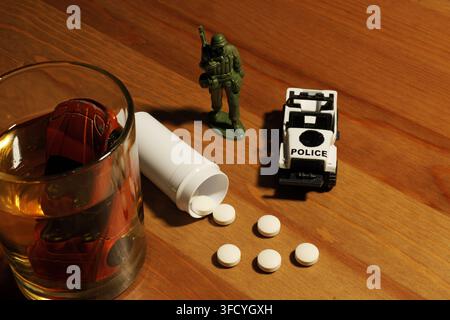 Kleines rotes Auto-Spielzeug, das in einem Glas Whiskey versinkt, neben verschütteten weißen Pillen aus einer Flasche, einem Spielzeugsoldaten und einem Spielzeug-Polizeiauto auf einem Holztisch, depi Stockfoto