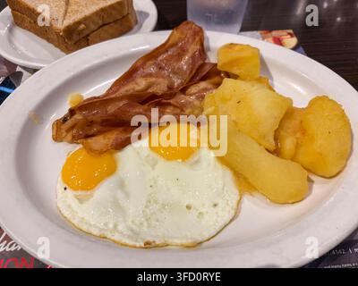 Zwei Eier mit Speck und Kartoffel-Frühstück auf einem weißen Teller, serviert an einem Restauranttisch mit Toast. Stockfoto
