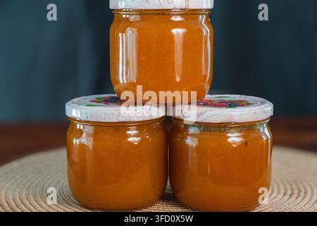 Ein Stapel von drei Gläsern, gefüllt mit hausgemachter Orangenmarmelade, steht auf einer runden gewebten Matte vor einem dunklen Hintergrund. Stockfoto