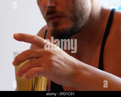 Detaillierte Nahaufnahme einer Hand eines Mannes, die ein hohes Glas mit einem kalten gelben Eiskelch und einem Strohhalm hält. Der untere Teil seines bärtigen Gesichts ist sichtbar Stockfoto