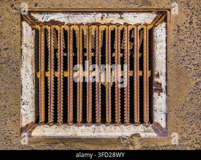 Nahaufnahme eines rostigen Metall-Sturmdrain-Gitters, das in strukturierten Beton eingelassen ist. Das Gitter zeigt erheblichen Rost und Schmutz und unterstreicht den urbanen Verfall. Stockfoto