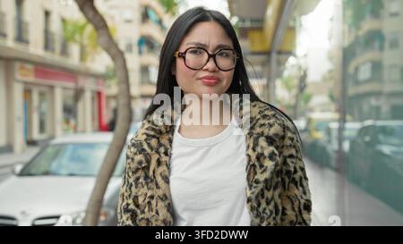 Frau im Leopardenmantel zuckt mit Handflächen auf der Straße, während sie den Kopf neigt, Brille und weißes T-Shirt trägt; Unklarheit. Stockfoto
