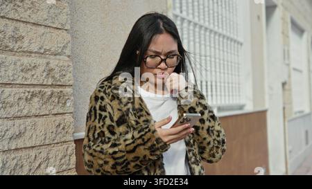 Frau hält Smartphone und tippt auf den Bildschirm, trägt Brille und Leopardenfellmantel, steht an einer Steinmauer und einem Metallgitter auf der Straße; ruhige Besorgnis. Stockfoto