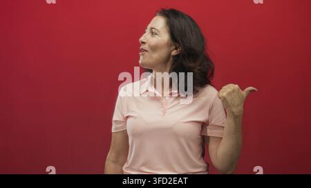 Mitteldalte Frau lächelt nach rechts und zeigt den Daumen nach hinten, während sie ein pinkes Polohemd gegen roten Hintergrund im Studio trägt; Selbstvertrauen. Stockfoto