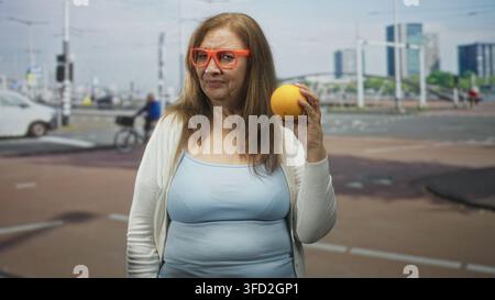 Frau mit Orange in der rechten Hand, orange Brille und hellblaues Tanktop auf der Straße; spielerische Neugier. Stockfoto