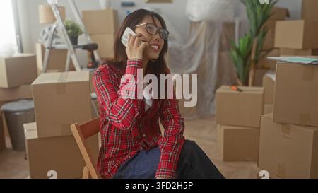 Frau, die auf einem Stuhl sitzt und Telefon an Ohr hält, zwischen Umzugskartons aus Pappe, Leiter und Topfpflanze im Gebäude; Erwartung des bewegenden Tages. Stockfoto