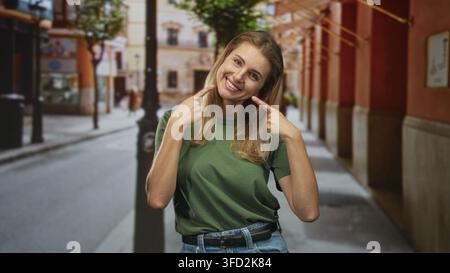 Frau zeigt Zeigefinger auf lächelnde Zähne auf einer sonnigen Straße mit Efeu und Schaufensterfronten; fröhlicher ehrlicher Moment. Stockfoto