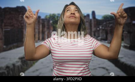 Die Frau zeigt mit den Zeigefingern nach oben auf die Steinstraße, trägt ein rosa gestreiftes Hemd und blickt nach oben; Reisewunderung. Stockfoto