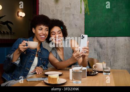 Zwei schöne junge Frauen genießen Kaffee und Lachen zusammen, während sie in einem stilvollen Café Selfies machen. Stockfoto