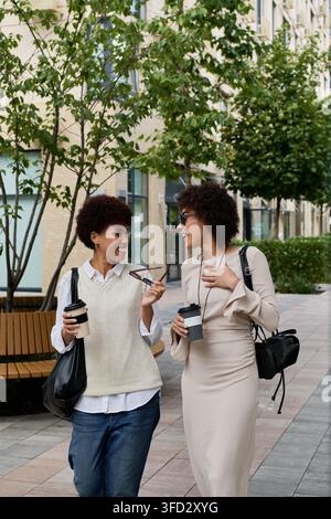 Zwei stilvolle Freunde teilen Gelächter und Kaffee, während sie durch einen lebendigen Stadtplatz schlendern. Stockfoto